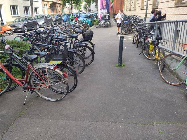 Foto vor der Schule mit auf dem Bügrersteig abgestellten Fahrrädern sowie Fahrräder in Fahrradständern